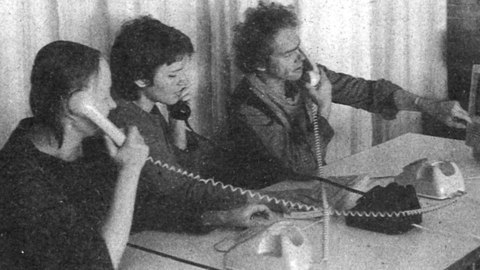 Schwarz-weiss Foto mit zwei Frauen und einem mann, die nebeneinander sitzen und telefonieren.