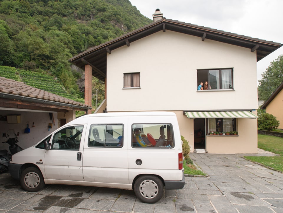 Sofia und Enea blicken aus dem Fenster im ersten Stock vom Haus der Familie Leonardi. Vor dem Haus steht das Familienauto, ein weisser Van.