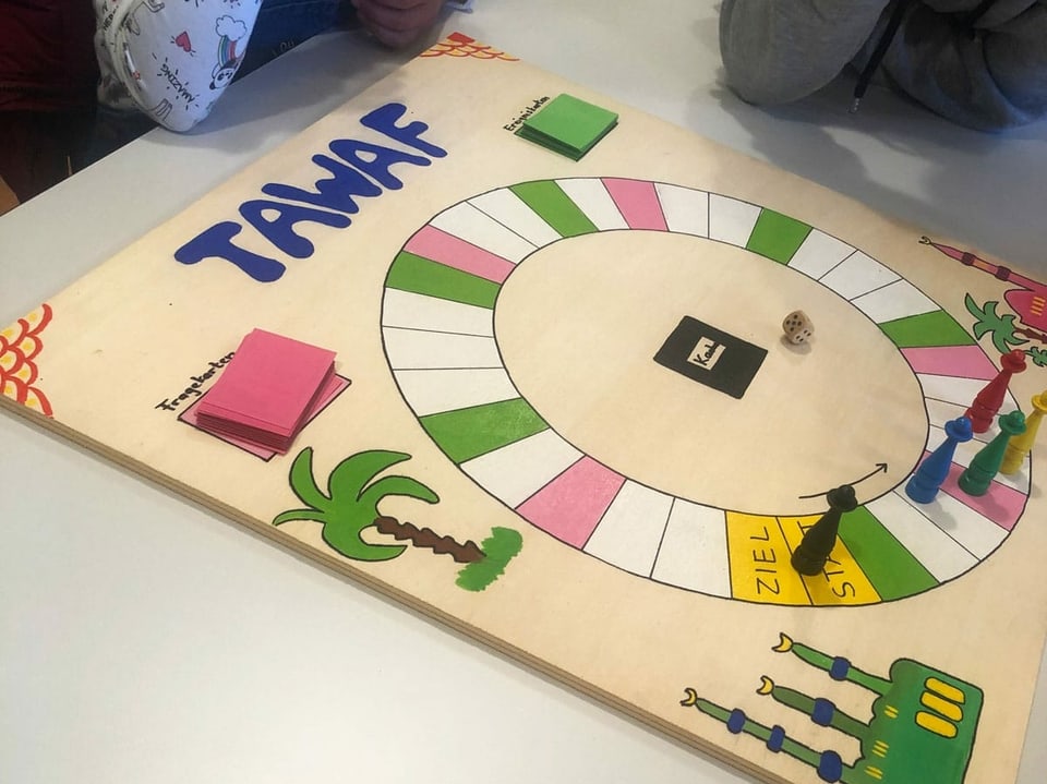 Ein quadratisches Spielbrett mit zahlreichen weissen, grünen und pinkfarbenen Feldern, die im Kreis angeordnet sind, liegt auf einem Tisch.