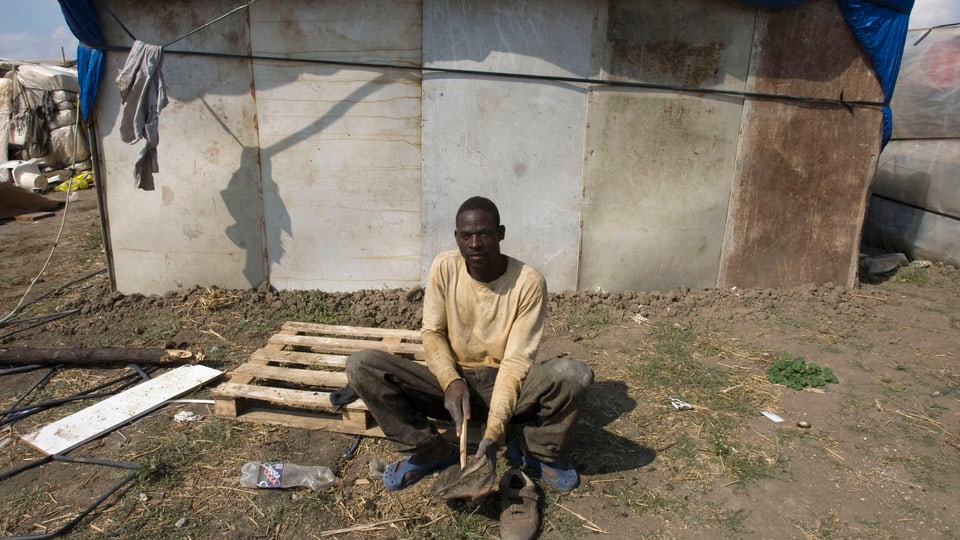 Ein afrikanischer Mann sitzt vor einer baufälligen Hütte auf einem Feld.