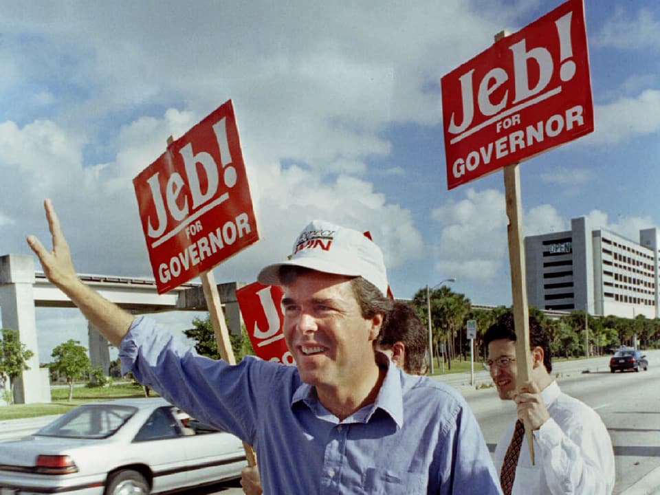 Jeb Bush steht mit Anhängern neben einer Strasse und hält Wahlplakate in den Händen.