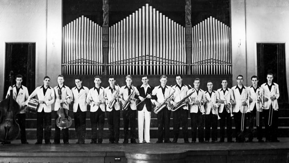 Männer in weissen Blazer halten Instrumente und stehen in einer Reihe auf der Bühne.