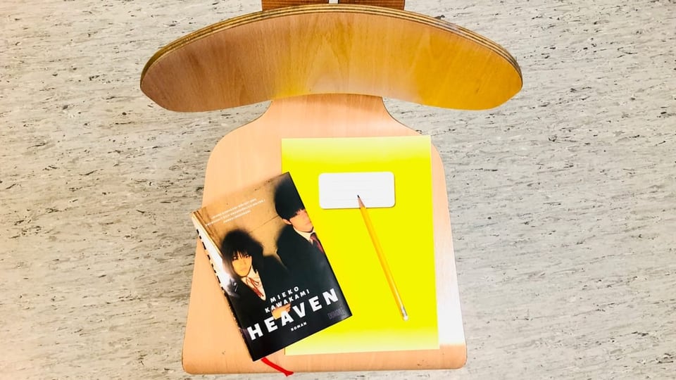 «Heaven» von Mieko Kawakami liegt zusammen mit einem Schulheft und einem gelben Bleistift der Stärke 6B auf einem Stuhl.