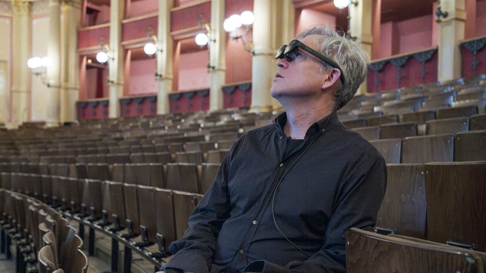 Ein Mann mit schwarzen Hemd sitzt in den leeren Sitzreihen eines Theaters in Bayreuth. Auf dem Kopf eine AR-Brille