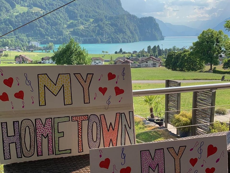 Ein «My Hometown»-Plakat steht links unten im Bild auf einem Balkon. Im Hintergrund sieht man einen blauen See und Berge