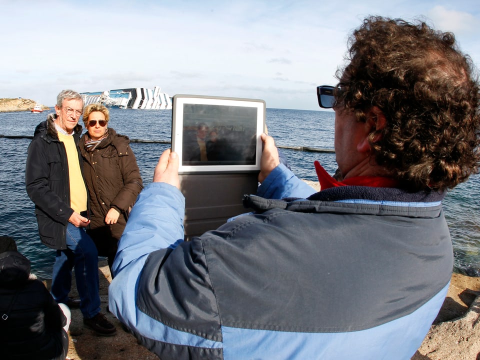 Ein Mann fotografiert ein Paar. Im Hintergrund ist die Costa Concordia zu sehen.