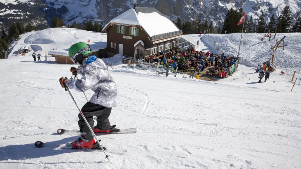 Kind auf Skiern auf einer Skipiste, im Hintergrund ein Restaurant mit voller Sonnenterrasse.