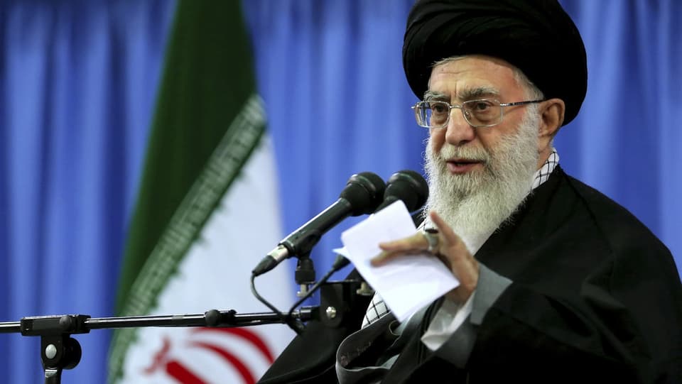 Khamenei mit schwarzem Turban spricht in ein Mikrofon, hinter ihm blaue Vorhänge.