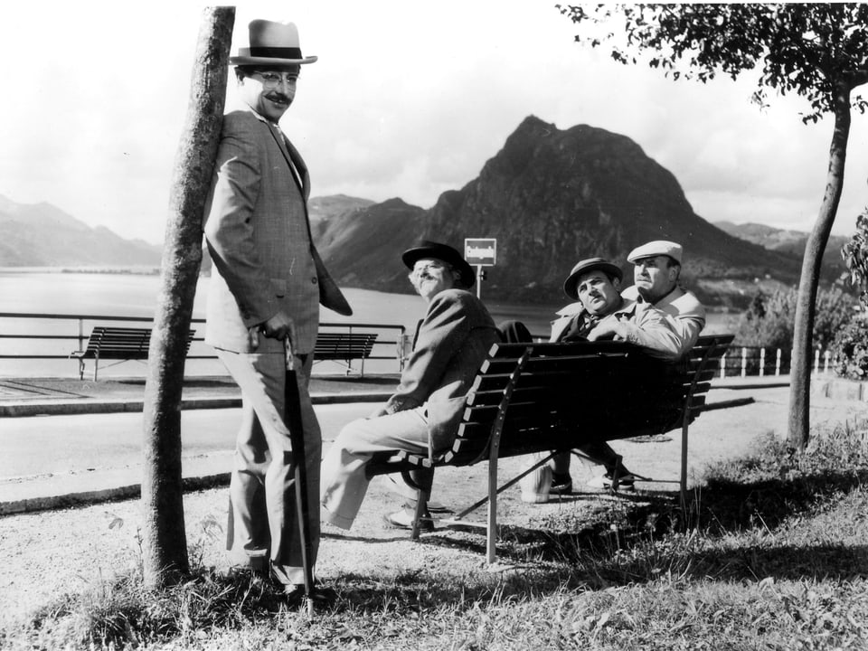  Drei Männer sitzen an auf einer Bank am Ufer eines Sees und blicken zu einem vierten Mann, der an einem Baum lehnt.