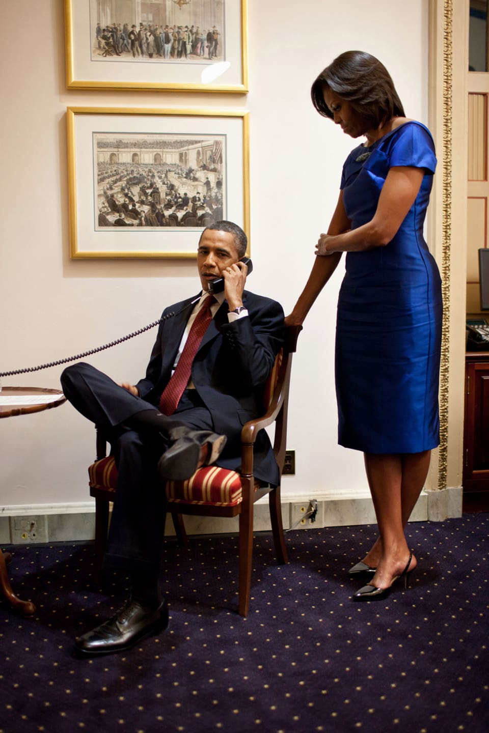 Barack Obama sitzend am Telefon. Neben ihm steh Michelle Obama im blauen Kleid.