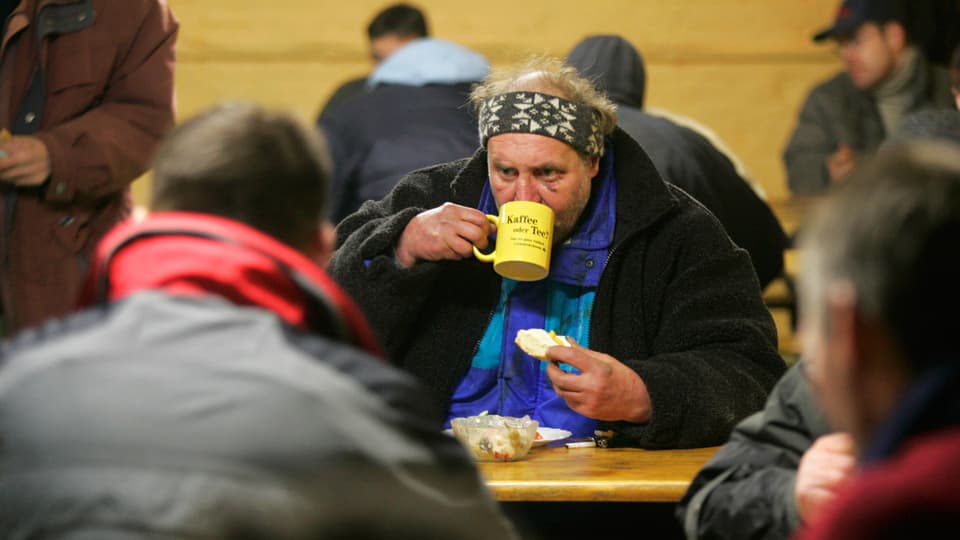 Ein obdachloser Manhn trinkt an einem Tisch aus einer Tasse, umgeben von anderen Menschen.