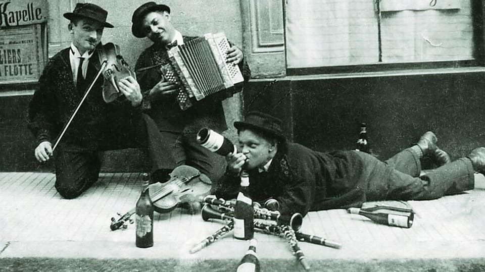Der Klarinettist Kasi Geisser liegt auf der Strasse am Boden und trinkt Wein mit seinen Kollegen.