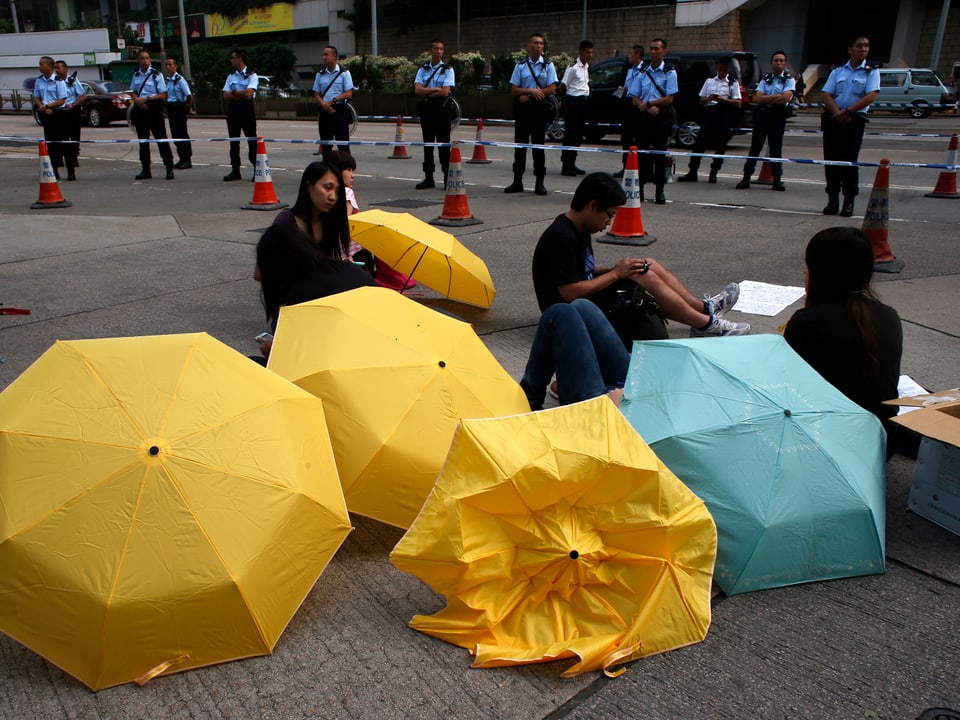 Drei Aktivistinnen sitzen hinter Regenschirmen auf der Strasse und werden von einer Polizeieinheit hinter einer Abschrankung beobachtet.