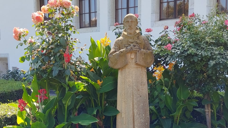 Statue eines Mönchs umgeben von bunten Blumen vor einem Gebäude.