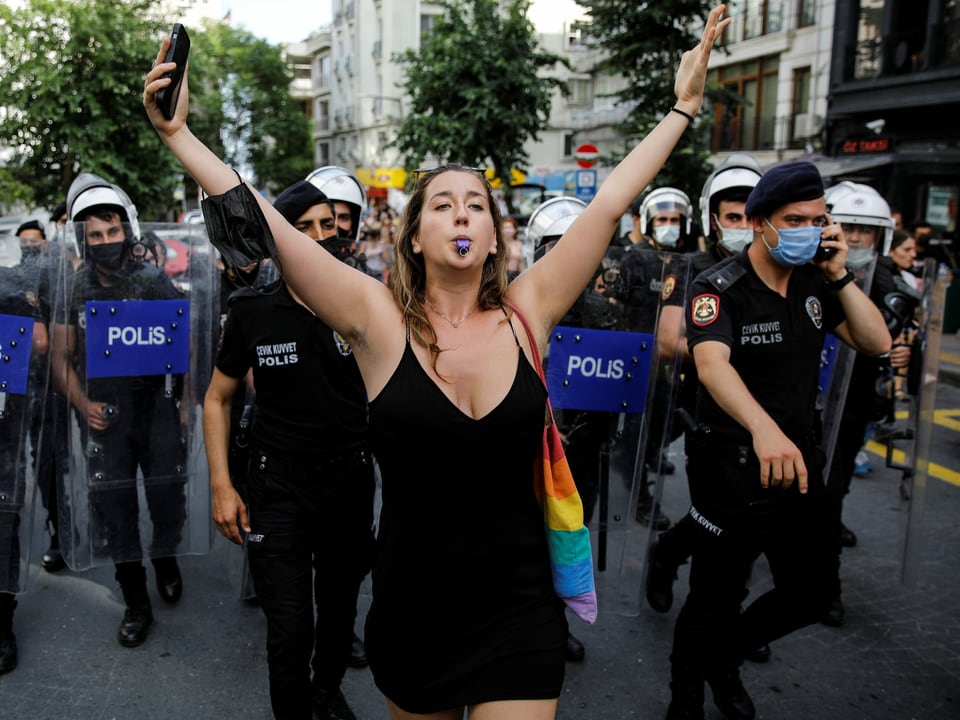 Eine Demonstrantin mit Regenbogentasche breitet die Arme aus und pfeift mit einer Trillerpfeife, hinter ihr Polizei