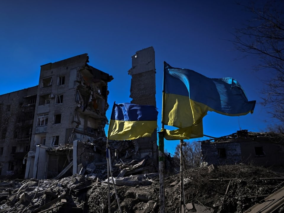 Ukrainische Nationalflaggen wehen vor einem schwer beschädigten Wohnhaus.