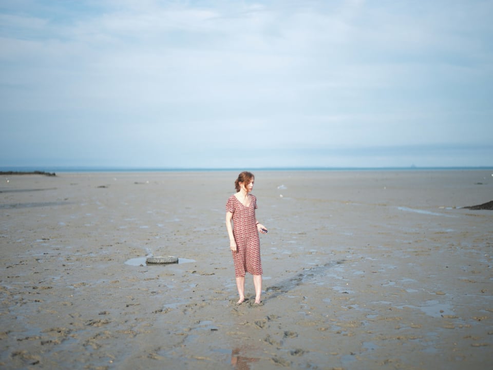Eine Frau steht barfuss am Strand und schaut wirr umher.