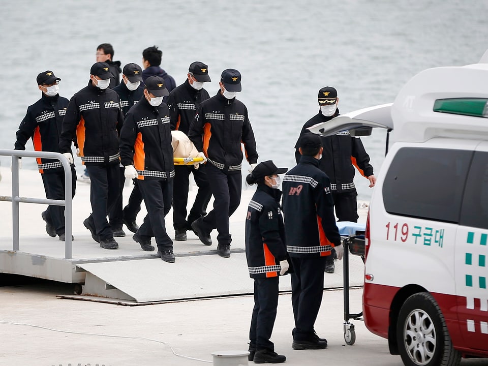 Ein Team in Uniform und mit Schutzmasken trägt eine Bahre mit einer Leiche in einer weissen Hülle zu einer Ambulanz.
