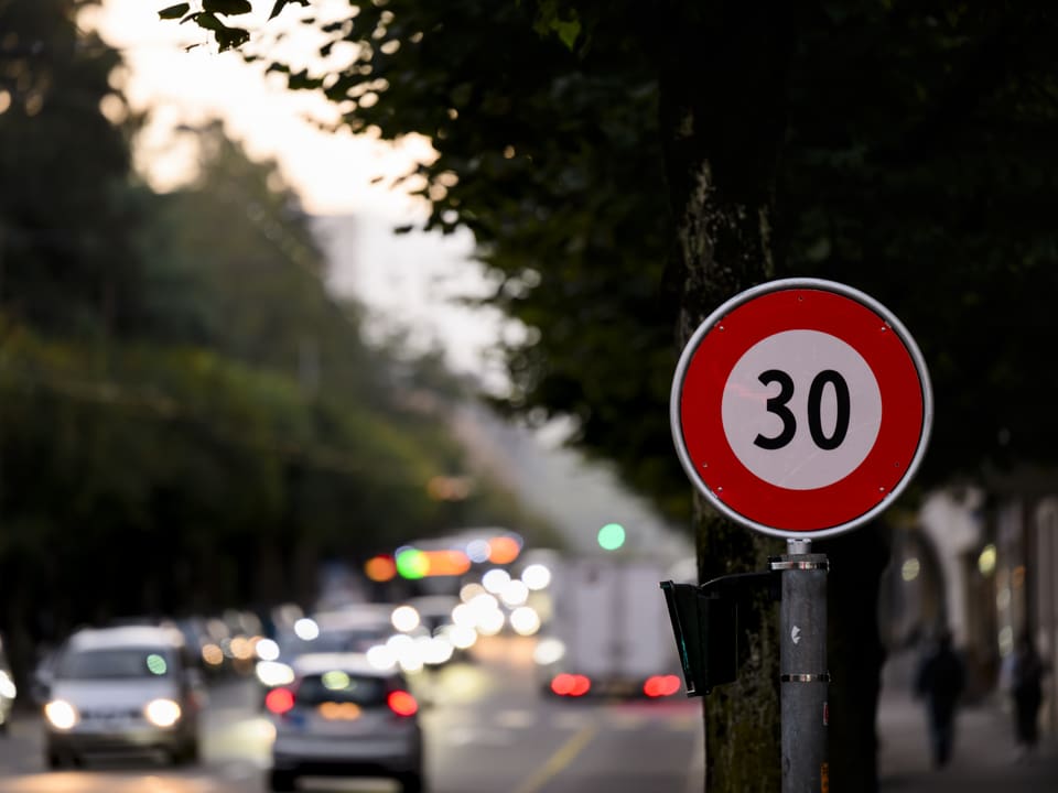 Verkehrsschild mit Geschwindigkeitsbegrenzung 30 vor unscharfem Strassenhintergrund bei Dämmerung.