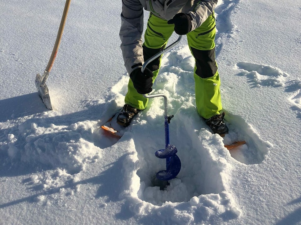 Ein Mann bohrt ein Loch ins Eis auf einem Bergsee.