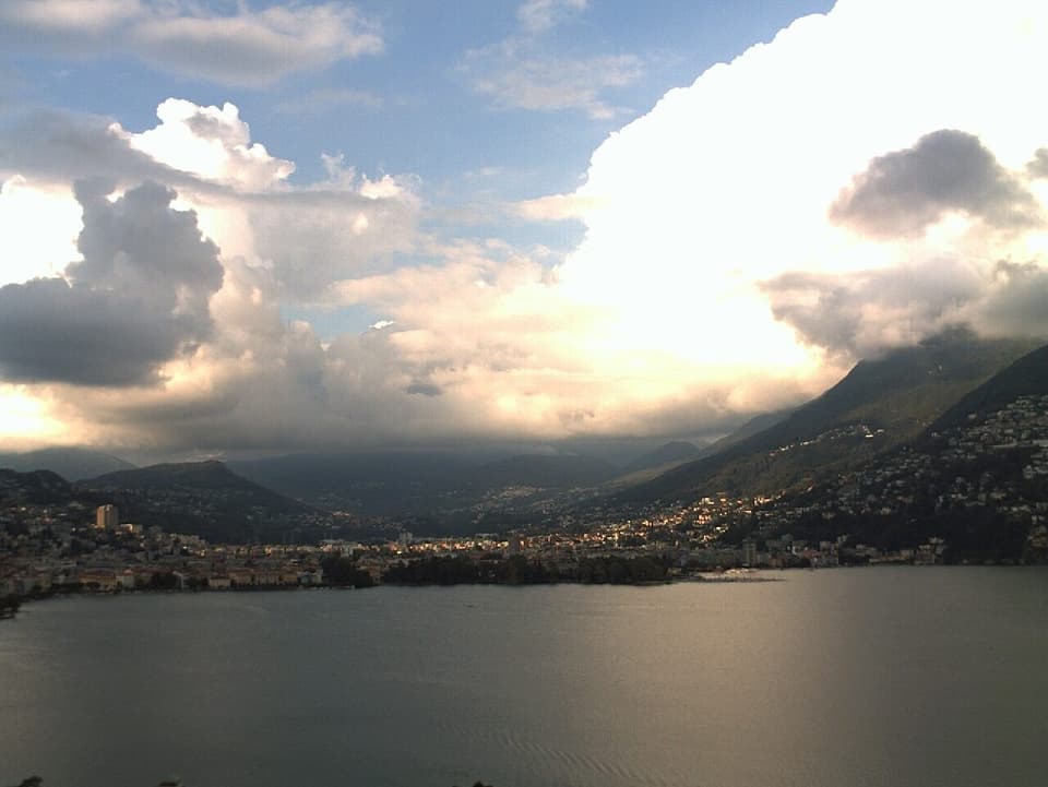 Im Vordergrund Lugano und der Luganersee. Im Hintergrund werden die grossen Quellwolken an den Hügel von der Abendsonne angestrahlt.