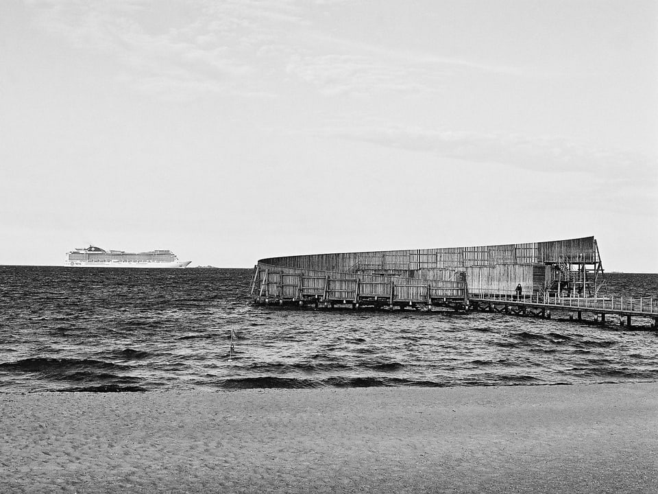 Eine hölzerne Badeanstalt an der Meeresküste. Im Hintergrund ein Kreuzfahrtschiff (Schwarz-Weiss-Bild)