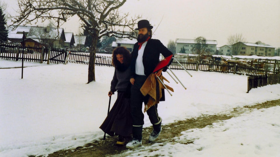 Röbi mit Hut spaziert mit seiner Frau Heidi, die einen Gehstock in der rechten Hand hält, im Schnee.