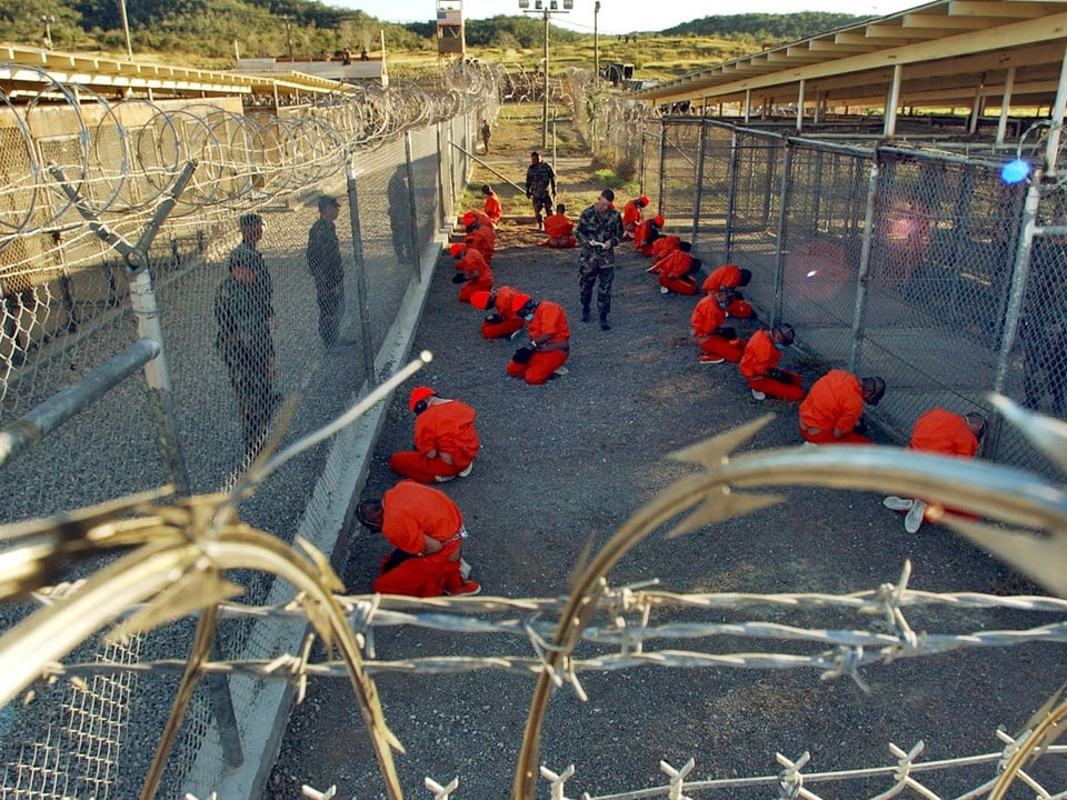 Zahlreiche Gefangene in orangen Overalls knien auf dem Erdboden.