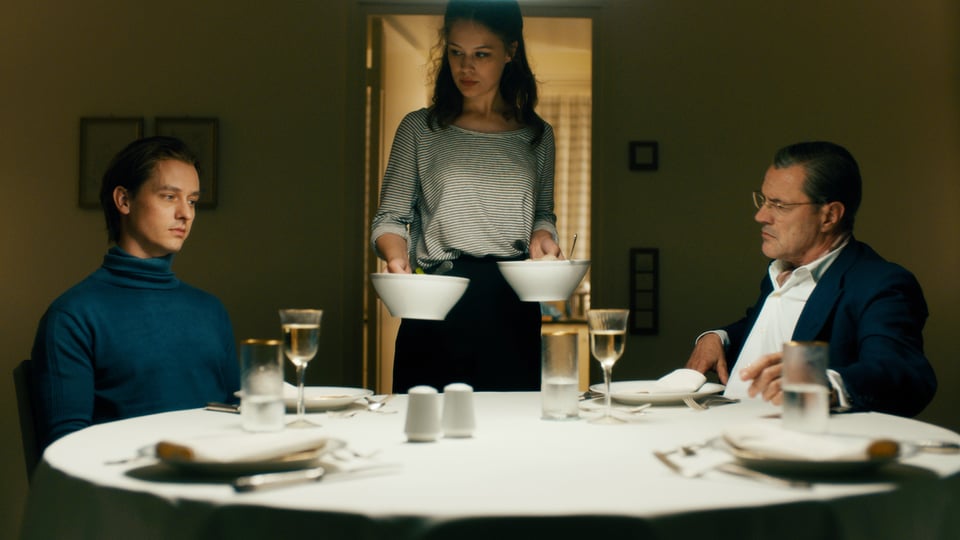 Zwei Männer am Tisch, eine Frau steht mit Tellern in der Hand daneben.