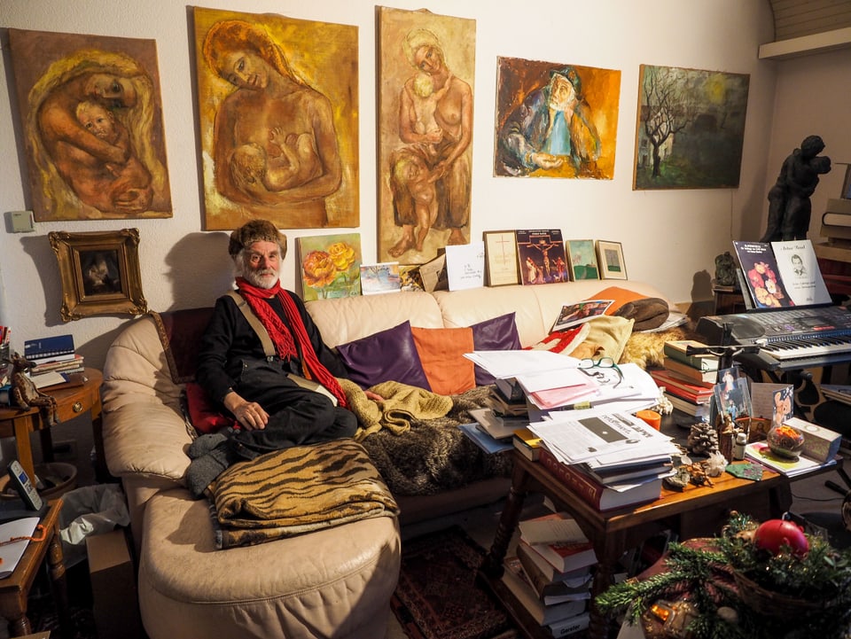Ernst Sieber sitzt vor einer Bilder-Wand auf einem Sofa.