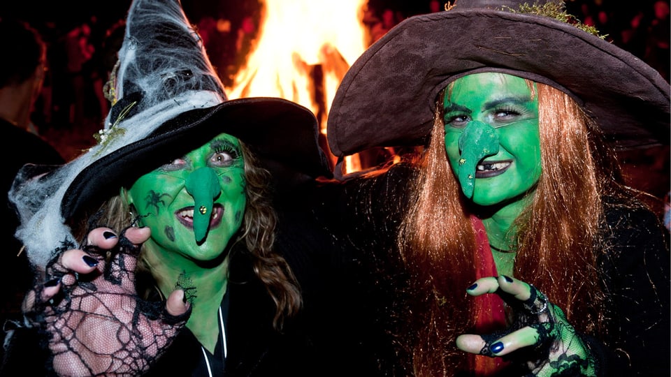 Zwei Hexen mit grünen Gesichtern und krummen Nasen, hinter ihnen lodert ein Feuer.