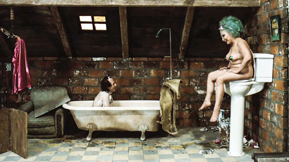 Altes Badezimmer: Ein Mann sitzt in der Wanne und lacht eine nackte Frau mit grünem Haar an, die auf einer erhöhten Toilette sitzt. 