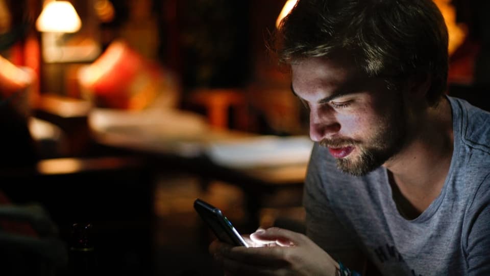 Mann blickt in dunklem Wohnzimmer auf sein Smartphone.