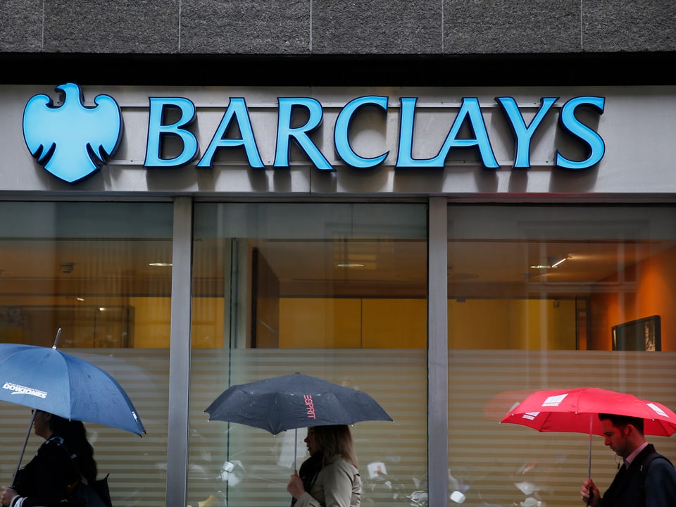Personen mit Regenschirmen laufen an einer Barclays-Bankfiliale vorbei.