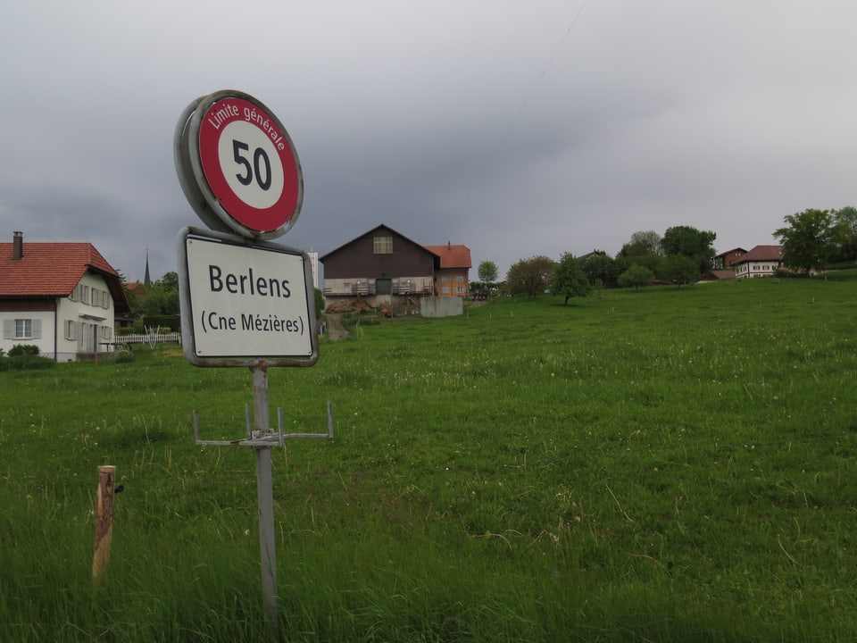 Ein Ortschild mit der Aufschrift Berlens vor einer grünen Wiese, im Hintergrund ein Bauernhof.