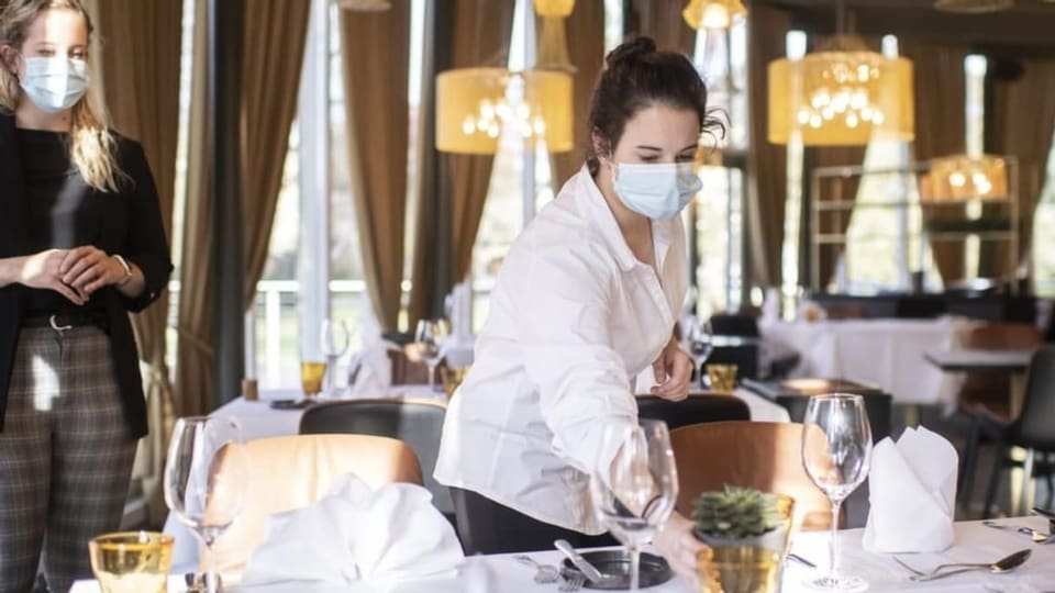 Eine Angestellte mit Hygienemaske deckt einen Tisch, ihre Ausbildnerin schaut im Hintergrund zu.