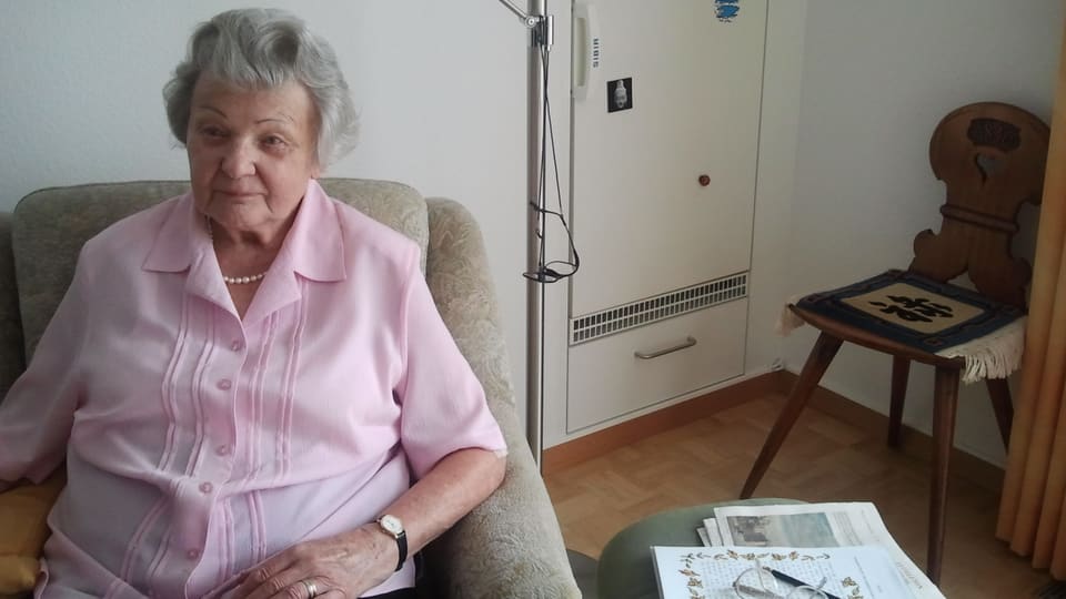 Die Seniorin mit roas Bluse sitzt in einem bequemen Sessel in ihrem Wohnzimmer.