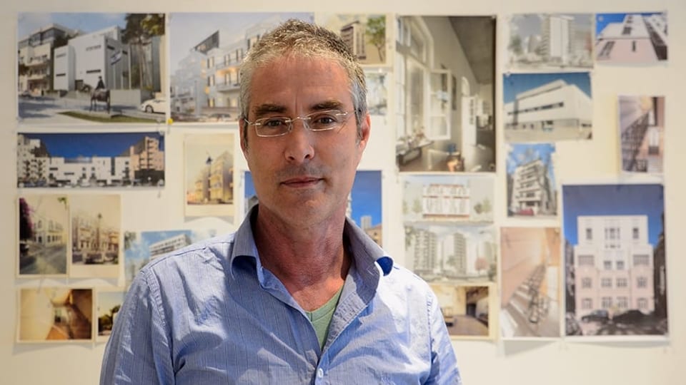 Ein mann mit hellblauem Hemd, hinter ihm an der Wand hängen Fotos von Gebäuden.