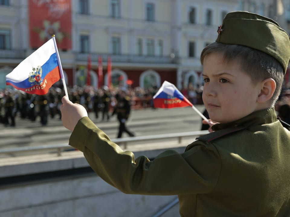 Ein kleiner Junge in Uniform schwingt die russische Fahne, während er die Parade verfolgt.