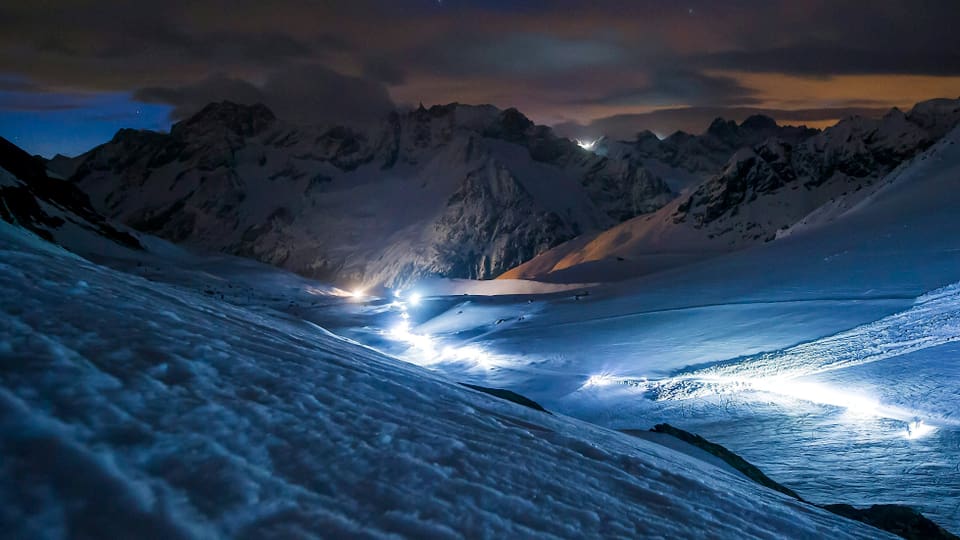 Offenes Hochtal mit Leuchtspur der Sportler. Im Hintergrund schorffe Berge, im Vordergrung viel Schnee. Alles im diffusen Licht der Nacht. 