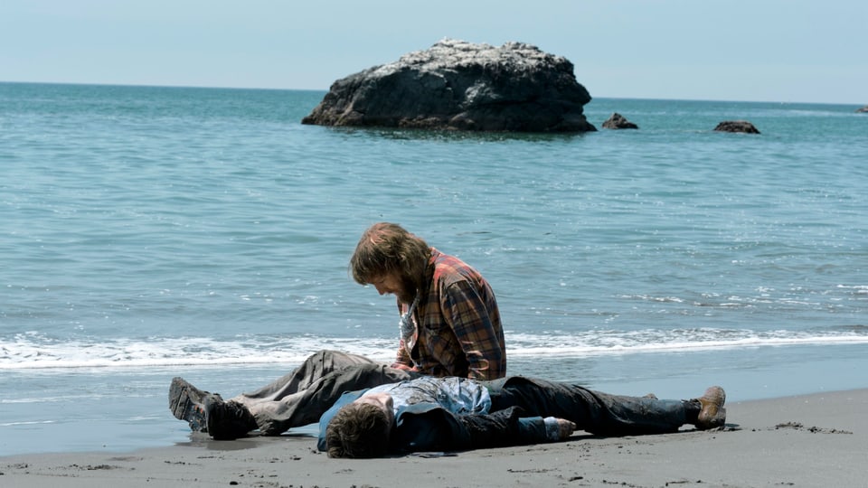 An einem Strand. Ein Mann liegt im Sand, ein anderer Mann sitzt neben ihm und schaut auf den Boden