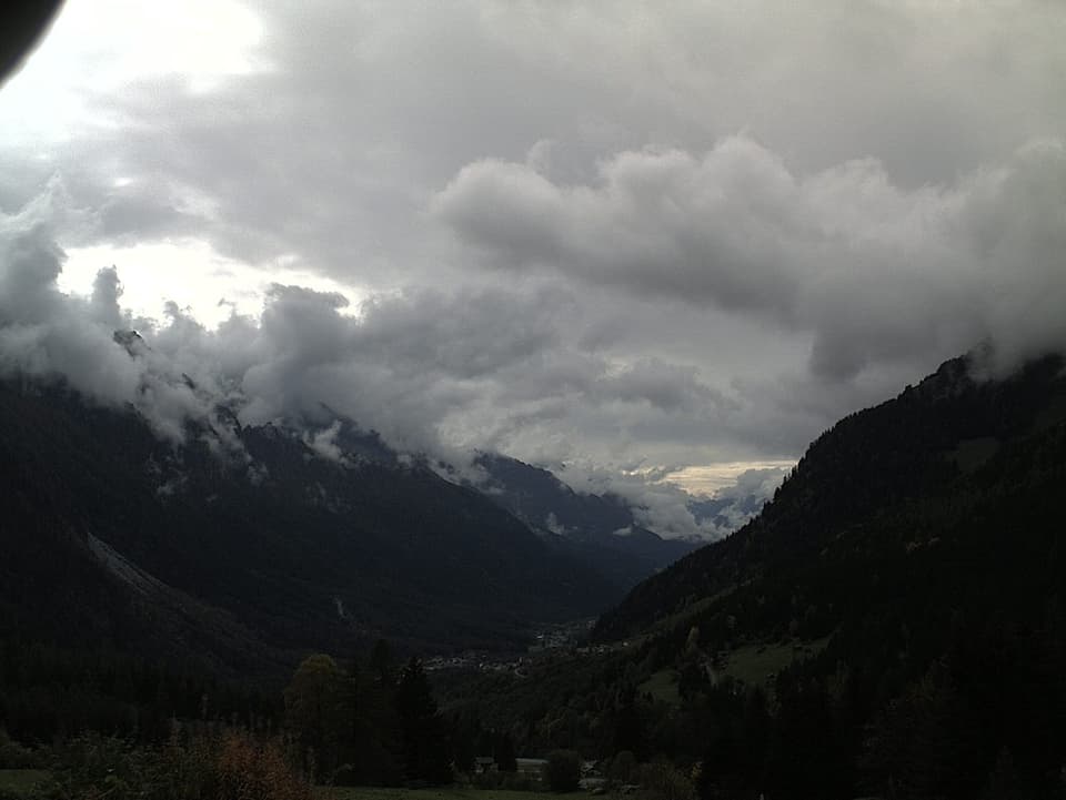 Das bewaldete Tal von Bergell erscheint hell und dunkel grün. An den oberen Hängen zeigt das Bild abstehende Wolkenfetzen in verschiedenen Grautönen. Der Himmel darüber ist ebenfalls verschieden grau.