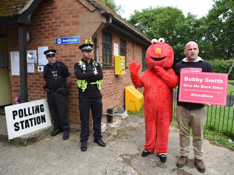 Verkleideter Mann in rotem Elmo-Kostüm und ein Wahlhelfer mit Schild stehen vor Wahl-Lokal, daneben zwei Polizisten.