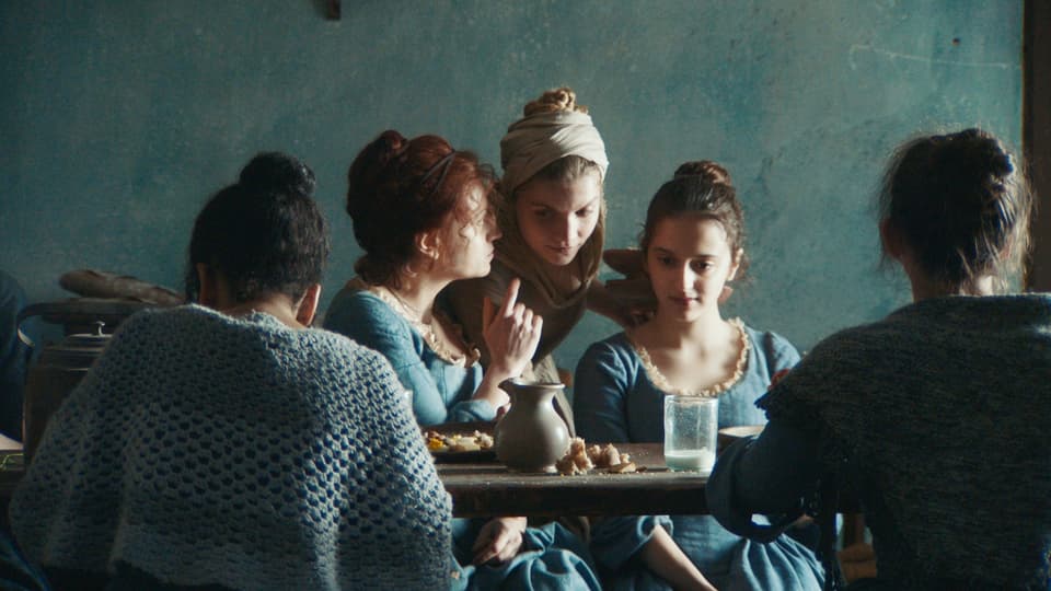 Mehrere Mädchen in barocken Bekleidung sitzen am Tisch: Eine flüstert der anderen etwas ins Ohr. 