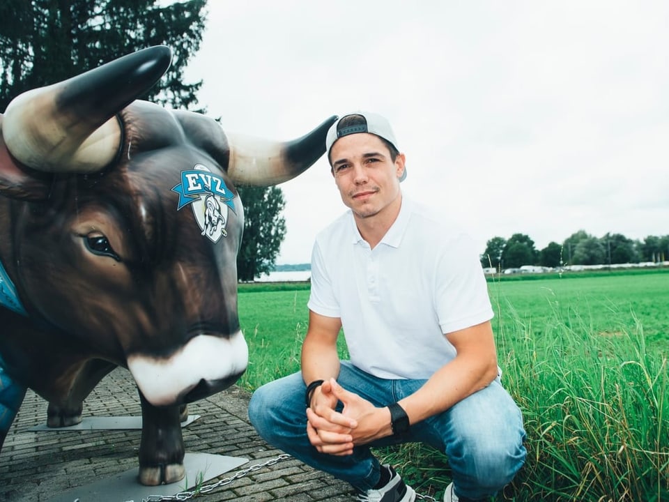 Ein Mann mit umgekehrter Dächlikappe sitzt neben einer Kuh-Statue.