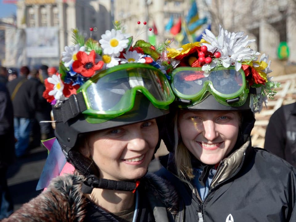 Zwei Mädchen mit Stahlhelm und Blumenschmuck lächeln für einen Fotografen.