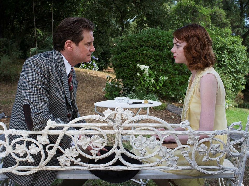 Colin Firth und Emma Stone schauen sich prüfend an. Zusammen sitzen sie an einem kleinen, weissen Tischchen in einem Park.