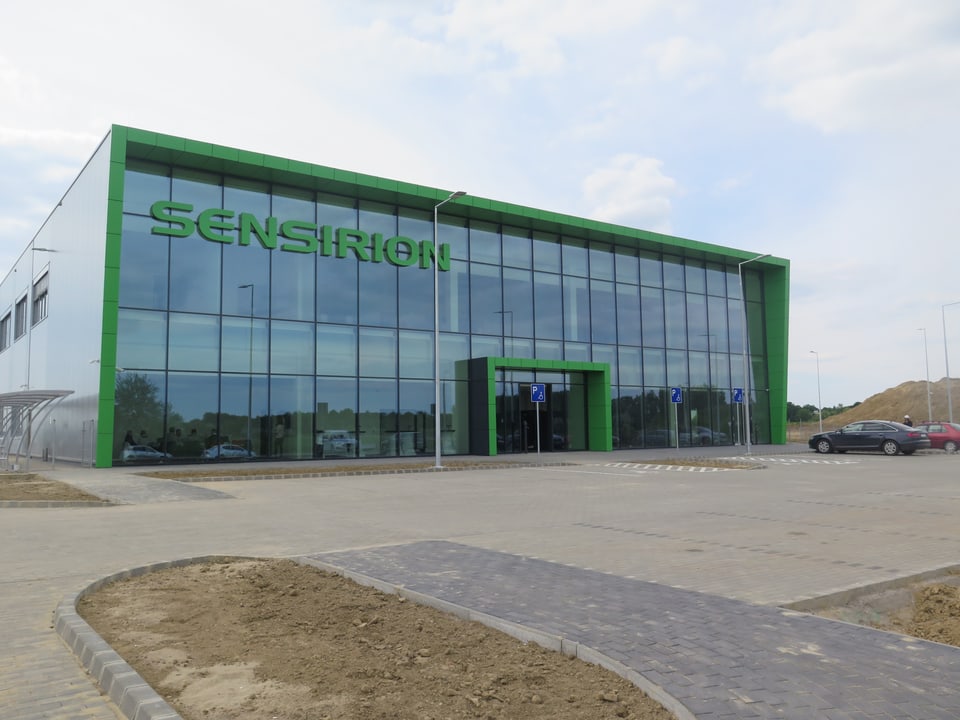 Ein modernes Fabrikgebäude mit grosser Glasfront. Das Logo ist grün. Davor ein Parkplatz.