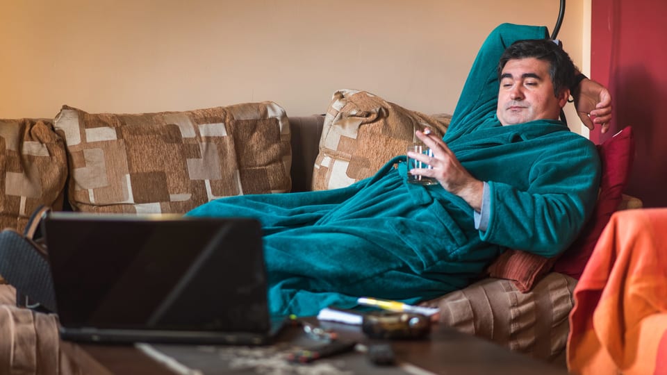 Ein Mann im Morgenmantel liegt auf dem Sofa, trinkt und schaut dabei in einen Laptop auf dem Sofatisch.