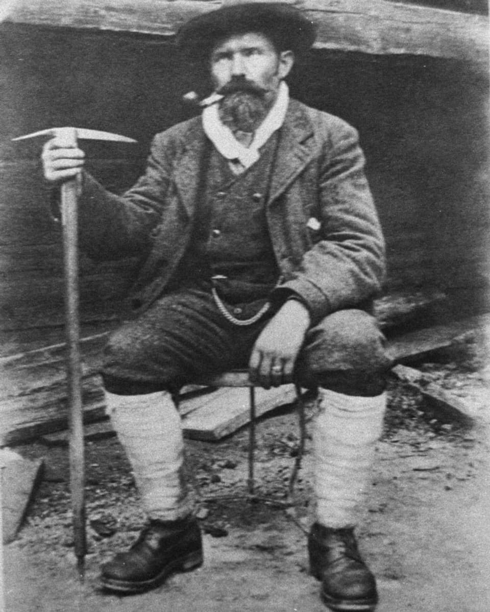 schwarzweiss-Bild eines Manns in Bergschuhen, mit Bickel ind er Hand, Hut auf und Pfeife im Mund.